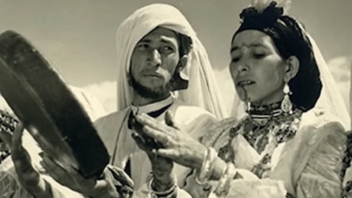 Black-and-white photograph of a woman with a chin tattoo and jewellery beside a man holding a frame drum