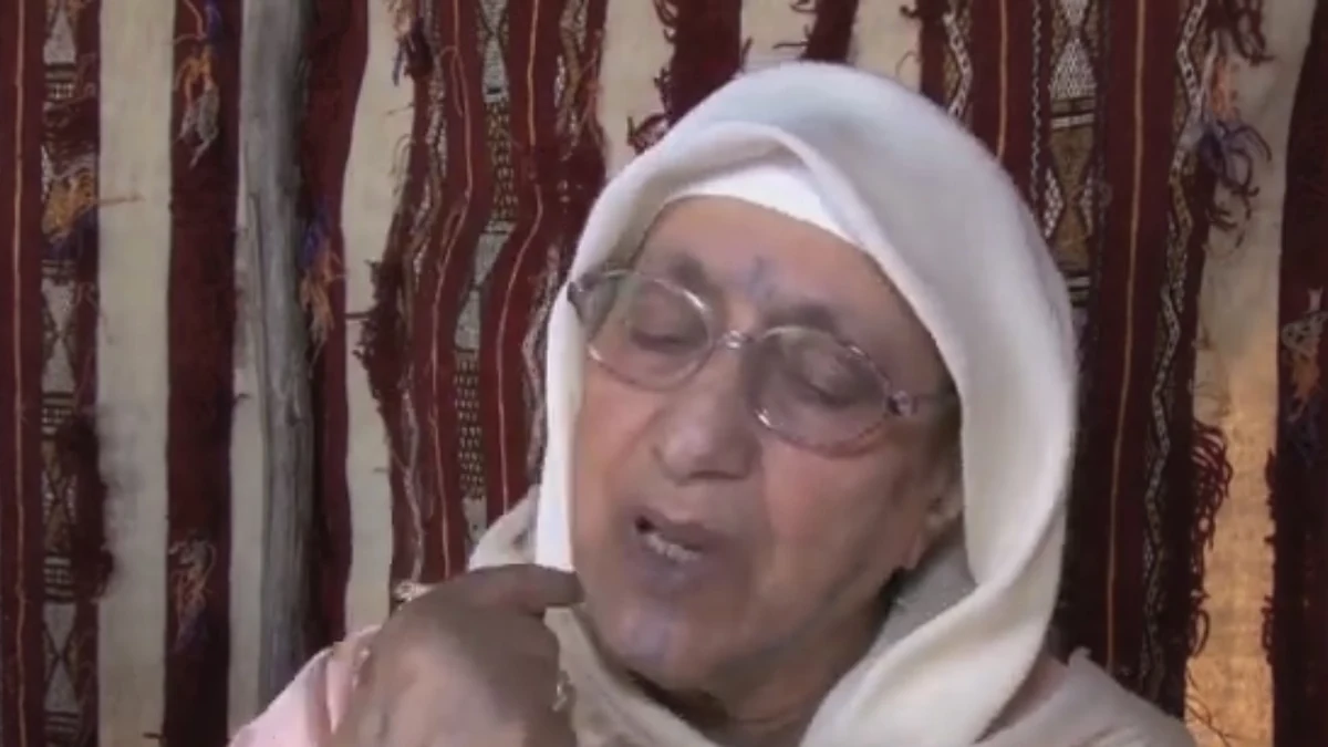 Woman wearing glasses and a white headscarf pointing towards her chin tattoo
